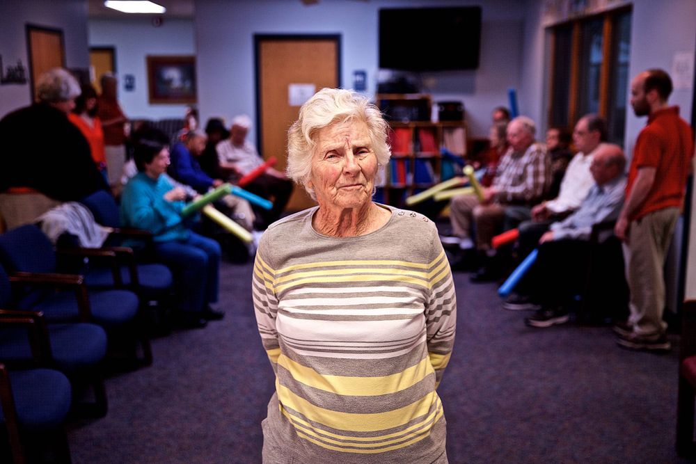 Photo of a woman standing looking into the camera with a serious expression, in an assisted living home, with other senior citizens in the background engaged in an activity. For a United Way advertising and collateral campaign. Photo of one of the many people that the United Way is assisting. Advertising photographer, on location photographer & location commercial photographer. Woman In a Assisted Living Facility