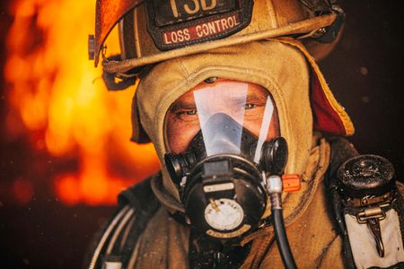 Shallow depth of field and selective focus. Very tight photo of Loss Control person dressed in protective fire gear, training and ready for action and to respond and mitigate a disaster as it relates to risk management in the insurance industry. The fire burns in the background. Environmental portrait, and we are a industrial photographer, on location photographer & location commercial photographer. Portrait of a Fireman