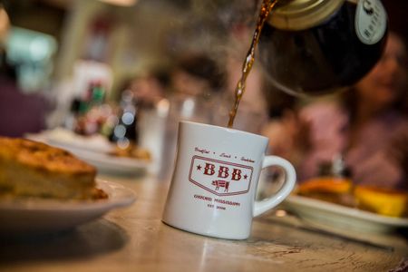 The coffee cup is the emphasis and focus as coffee is being pouted into, steaming hot. Photo at Big Bad Breakfast in Oxford, Mississippi. Shot for the Mississippi Tourism campaign for advertising, branding, collateral and broadcast motion assets. Environmental portrait, and we are a industrial photographer, on location photographer & location commercial photographer. Shallow depth of field and selective focus. VisitMS Coffee Pour at Big Bad Breakfast