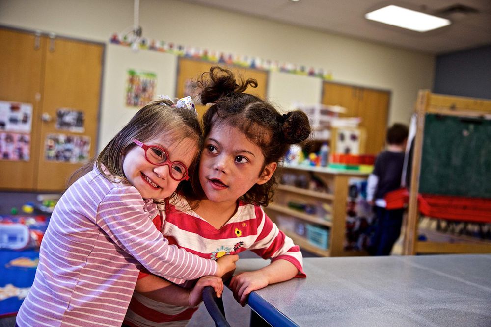 A photo a two young girls in a school classroom hugging and smiling, very affectionate, best friends, BFF. Photographed for a United Way advertising campaign. I am a advertising photographer, on location photographer & location commercial photographer. Photo of one of the many people that the United Way is assisting. Two Young Girls Hugging in School