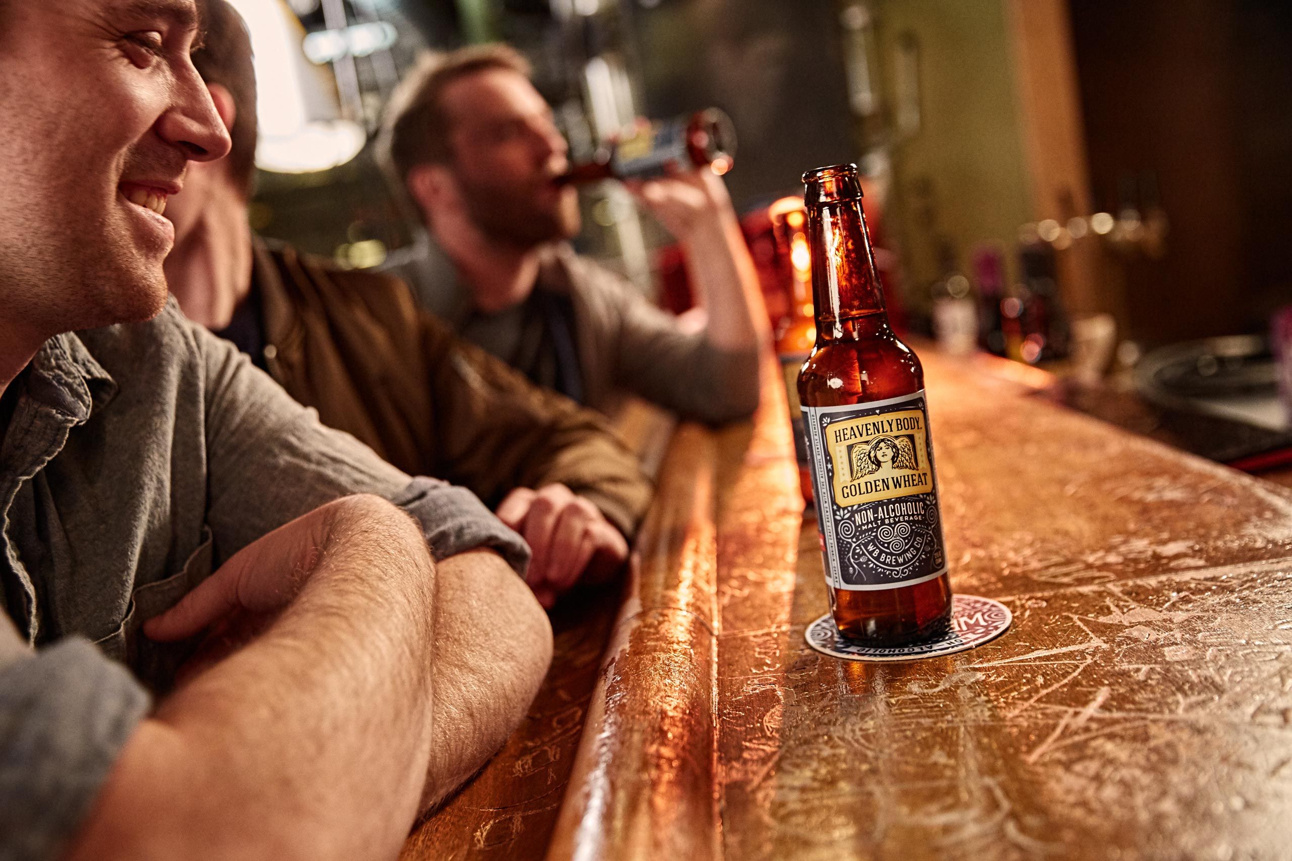 Long shot of a bar with WellBeing Heavily Body na non-alcoholic beers and a guy drinking one at the end. A smiling guy in the foreground and the drinker is out of focus. WellBeing Beers at a Bar