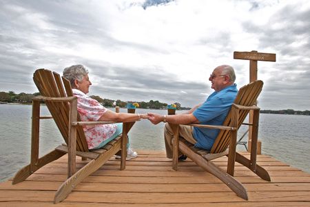 A retired couple, senior citizens sitting on a dock in Florida holding hands smiling and looking at each other with drinks on beautiful party cloudy sunny day. Retired Couple Sitting on a Dock
