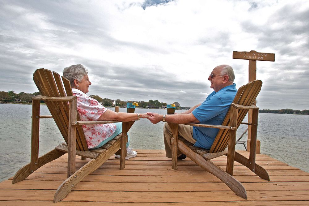 A retired couple, senior citizens sitting on a dock in Florida holding hands smiling and looking at each other with drinks on beautiful party cloudy sunny day. Retired Couple Sitting on a Dock