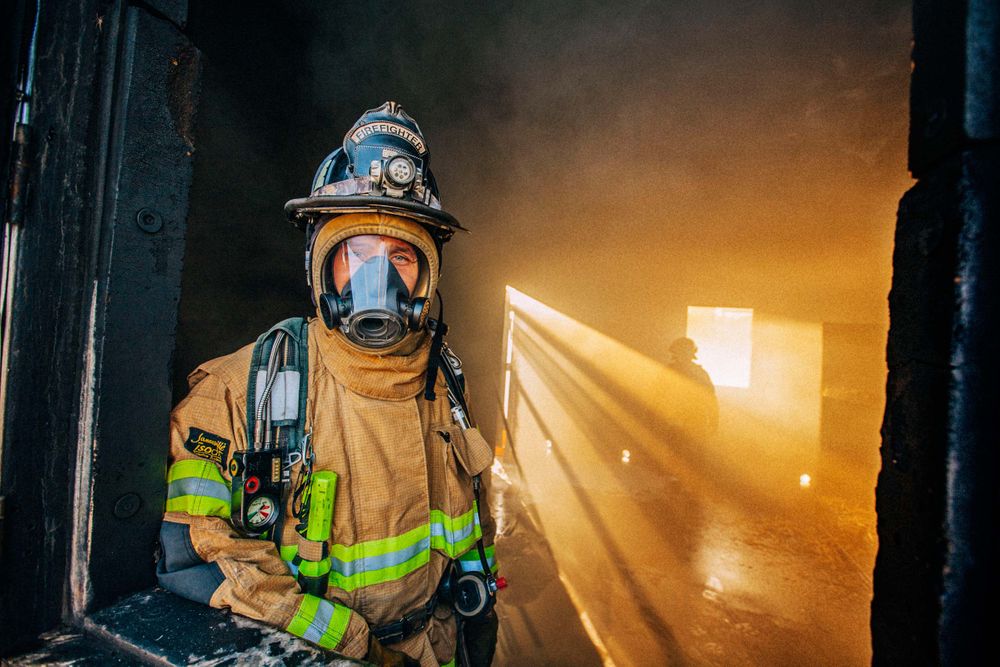 A fireman stands in a window of a burning and smoking building with fire & flames all around him, and really nice back light. Photo of Loss Control person dressed in protective fire gear, training and ready for action and to respond and mitigate a disaster as it relates to risk management in the insurance industry. Environmental portrait, and we are a industrial photographer, on location photographer & location commercial photographer. Fireman Looking Out of Smokey Window