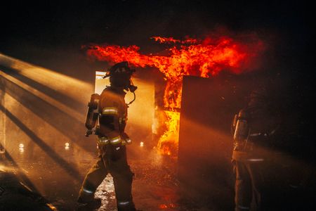 A fireman walks through a building with fire & flames all around him, and really nice light. Photo of Loss Control person dressed in protective fire gear, training and ready for action and to respond and mitigate a disaster as it relates to risk management in the insurance industry. Environmental portrait, and we are a industrial photographer, on location photographer & location commercial photographer. Firman Walks Through a Burning Building With Flames