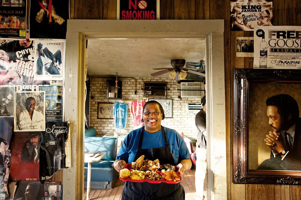 A photo of a woman holding a tray of food, in Bully's Restaurant in Jackson, MS, and smiling. Shot for the Mississippi Tourism campaign for advertising, branding, collateral and broadcast motion assets. Environmental portrait, and we are a industrial photographer, on location photographer & location commercial photographer. Shallow depth of field and selective focus. Woman Holding a Platter of Food in Bully's Restaurant