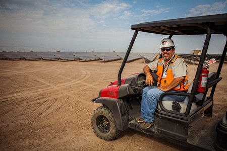 Environmental portrait of a worker. management type sitting on a Gator buggy on a dirt road with the solar field behind him. He smiling and is wearing protective glasses, helmet, vest etc. and wrapped and shielded from extreme heat, per OSHA requirement. This project was shooting a solar farm near El Centro, California, which is located right on the Mexico border. This energy collected from the sun will create free, off the grid, clean energy for many towns in California. As an industrial photographer, on location photographer & location commercial photographer this project was superb! Man On a Gator Buggy Smiling IN the Field