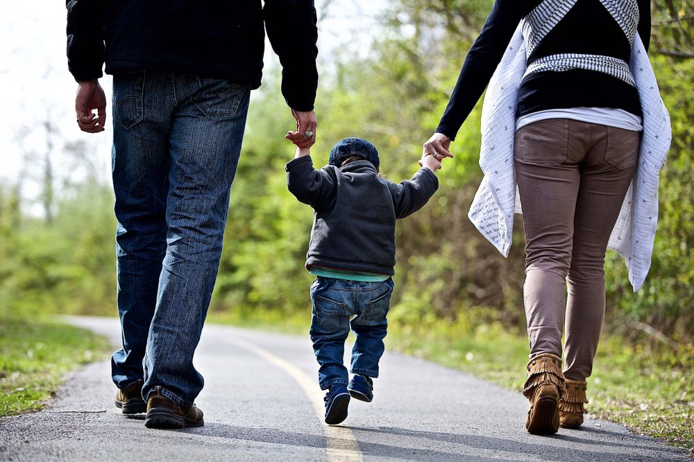 Photo of a family, photographed from behind, walking along a small path. The parents are holding hands with the your Son, in the middle and swinging him. Shot for a United Way advertising and collateral campaign. Photo of one of the many people that the United Way is assisting. Advertising photographer, on location photographer & location commercial photographer. Parents Walking With Their Child Photographed From Behind