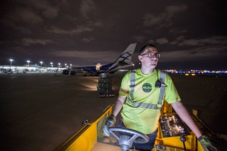 An cargo loader, worker, at an airport driving a yellow lift driving to drop more cargo. A Boeing 747-8 is seen in the background, at nighttime. He is wearing a protective vest and safety glasses. This was a project shooting for the air cargo transportation industry. This project was amazing as an industrial photographer, on location photographer & location commercial photographer. Shallow depth of field and selective focus with slow shutter speed and some motion blur. Airport Cargo Worker Driving a Lift
