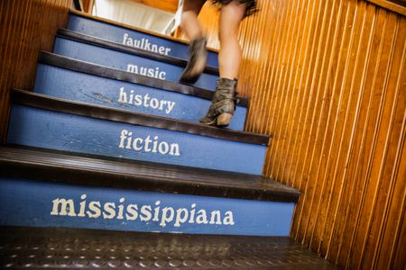 Photo a female walking up the stairs at Square Books in Oxford, MS. The stairs are painted blue and say Faulkner, Music, History, Fiction, Mississippiana. Shot for the Mississippi Tourism campaign for advertising, branding, collateral and broadcast motion assets. Environmental portrait, and we are a industrial photographer, on location photographer & location commercial photographer. Shallow depth of field and selective focus. VisitMS Employee Walking Up the Stairs at Square Books