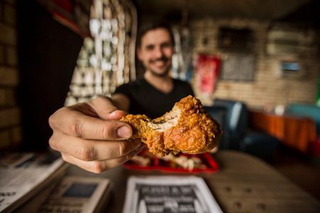 A photo of a man holding a piece of fried chicken, in Bully's Restaurant in Jackson, MS, toward the camera and smiling. the fried chicken is the emphasis and focus. Shot for the Mississippi Tourism campaign for advertising, branding, collateral and broadcast motion assets. Environmental portrait, and we are a industrial photographer, on location photographer & location commercial photographer. Shallow depth of field and selective focus. Man Holding Fried Chicken to the Camera