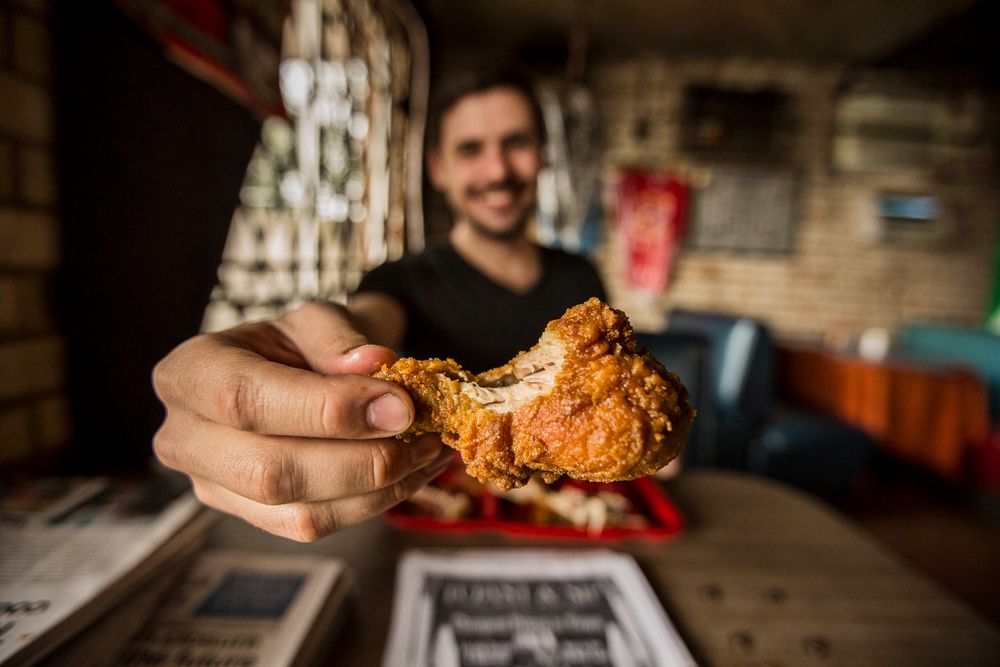 A photo of a man holding a piece of fried chicken, in Bully's Restaurant in Jackson, MS, toward the camera and smiling. the fried chicken is the emphasis and focus. Shot for the Mississippi Tourism campaign for advertising, branding, collateral and broadcast motion assets. Environmental portrait, and we are a industrial photographer, on location photographer & location commercial photographer. Shallow depth of field and selective focus. Man Holding Fried Chicken to the Camera