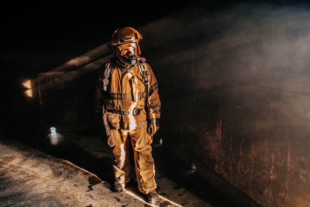 A fireman, full length, in a building surrounded with smoke, backlit with streaks of light, after the fire has been extinguished. Photo of Loss Control prevention person dressed in protective fire gear, training and ready for action and to respond and mitigate a disaster as it relates to risk management in the insurance industry. Environmental portrait, and we are a industrial photographer, on location photographer & location commercial photographer. Full Length of Firemen in Smoking Building