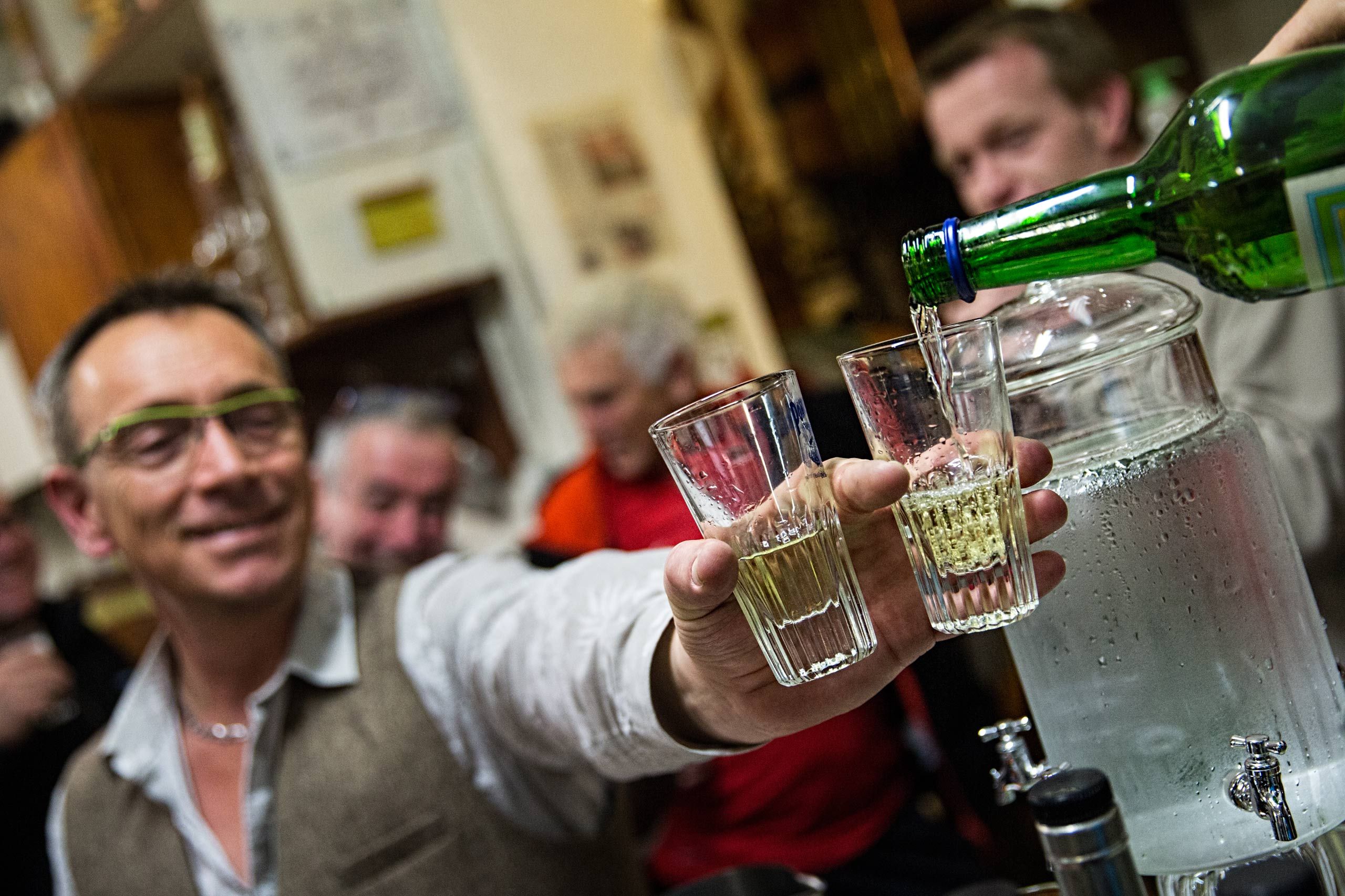 A photo of a man being served two glasses of absinthe at a party or gathering. Photos of the mysterious spirit, absinthe, also called wormwood, green fairy, green muse located at Distillery Armand GUY in Pontarlier, France. This was a passion project and an amazing adventure into the world of absinthe, especially as on location photographer & location commercial photographer. Photo was shot with a shallow depth of field and selective focus. The emphasis is on the glasses. Man Being Served Absinthe X2