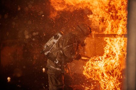 A fireman in a building surrounded with fire & flames all around him trying put them out. Photo of Loss Control prevention person dressed in protective fire gear, training and ready for action and to respond and mitigate a disaster as it relates to risk management in the insurance industry. Environmental portrait, and we are a industrial photographer, on location photographer & location commercial photographer. Fireman Putting Out Fire Surrounded By Flames