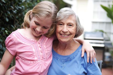 A smiling and happy Granddaughter in pink hugs her Grandmother dressed in blue outside on a nice day. Grandmother with Granddaughter