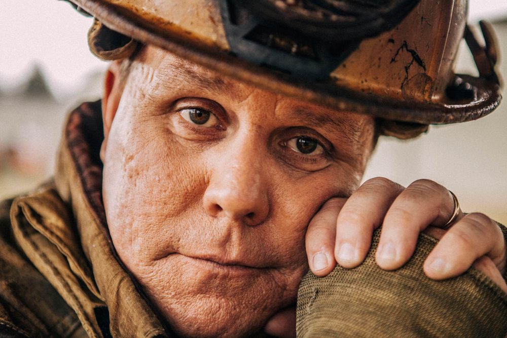 A fireman looks directly into the camera leaning his face on his hands. Photo of Loss Control person dressed in protective fire gear, training and ready for action and to respond and mitigate a disaster as it relates to risk management in the insurance industry. Environmental portrait, and we are a industrial photographer, on location photographer & location commercial photographer. Portrait of a Fireman