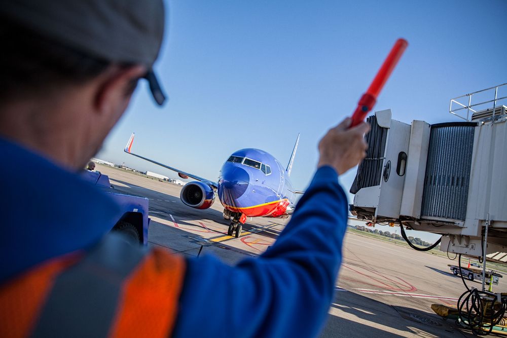 Photo of Southwest Airlines employee on the tarmac, marshaling a Boeing 737 the gate, at STL St. Louis Lambert International Airport FLYSTL Environmental portrait, and we are a industrial photographer, on location photographer & location commercial photographer. Shallow depth of field and selective focus. FLYSTL Aircraft Marshaling A Southwest Plane To the Gate