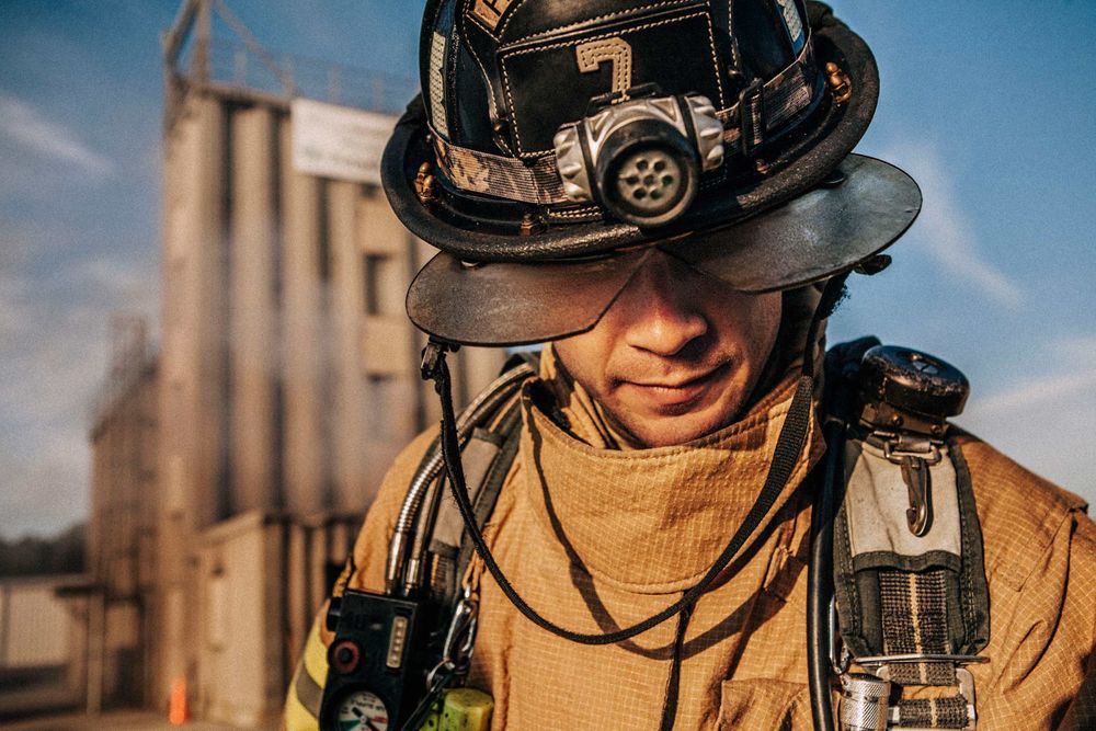 Photo of Loss Control person dressed in protective fire gear, training and ready for action and to respond and mitigate a disaster as it relates to risk management in the insurance industry. He is kneeling and preparing to enter a burin gin building. Environmental portrait, and we are a industrial photographer, on location photographer & location commercial photographer. Shot with Shallow depth of field and selective focus. Fireman Looking Down