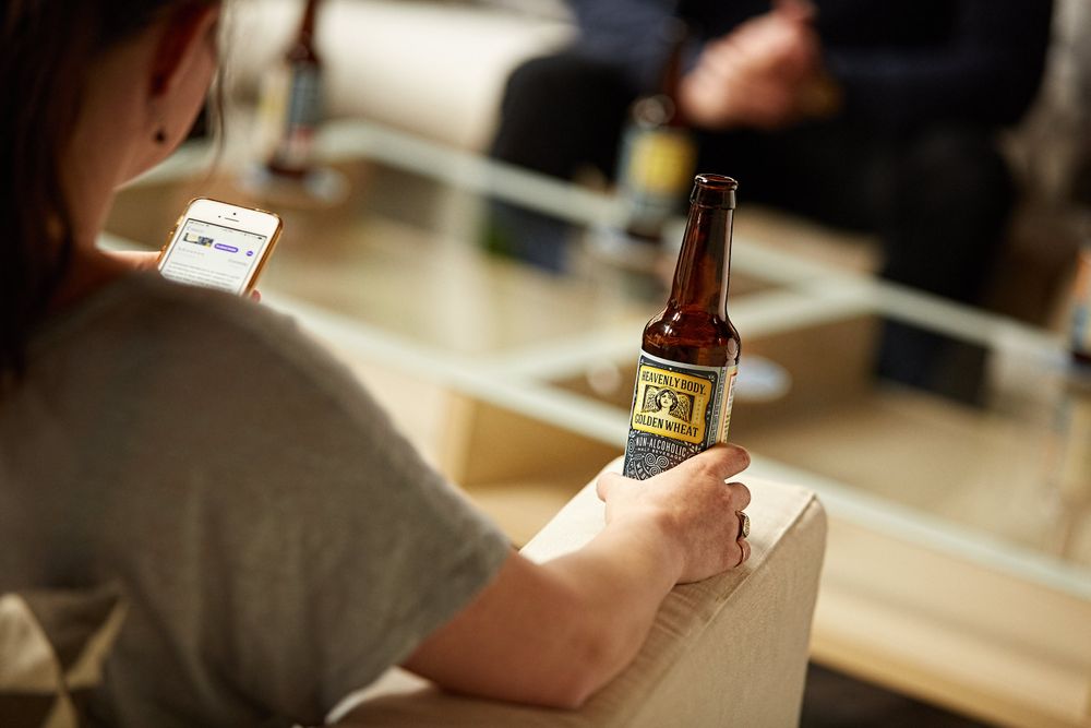 A woman sitting in the office during a meeting is texting and having a WellBeing na non-alcoholic beer. Woman Holding a Beer & Texting
