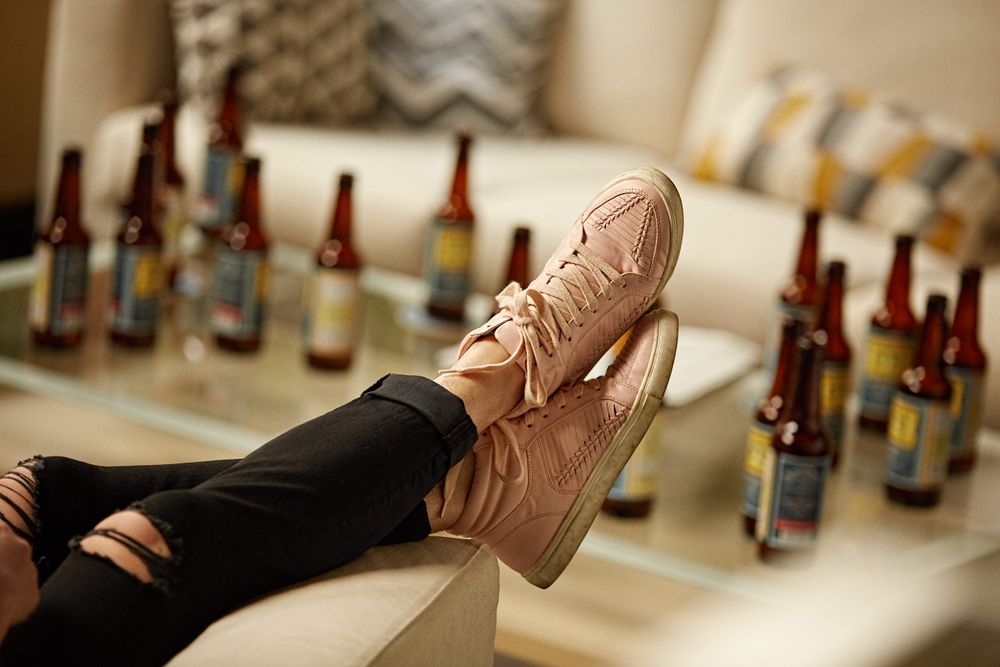 Women kicked back in a chair with her pink shows draped over the armrest and a table full of WellBeing Beer in the background out of focus. Emphasis is on the shows. Pink Shoes and Beer