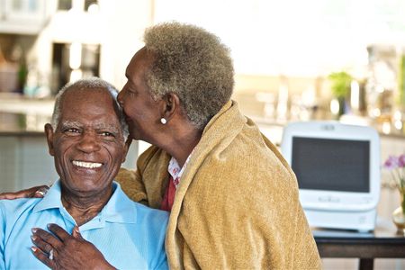 An African American senior citizen man smiling while being kissed by his wife with Intel health technology in the background. Senior citizen Couple Kissing and Smiling