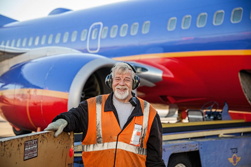 Photo of Southwest Airlines baggage handler on the tarmac, smiling & happy looking into the camera in front of a Boeing 737, parked at a gate, at STL St. Louis Lambert International Airport FLYSTL Environmental portrait, and we are a industrial photographer, on location photographer & location commercial photographer. Shallow depth of field and selective focus. FLYSTL Southwest Airline Baggage Handler