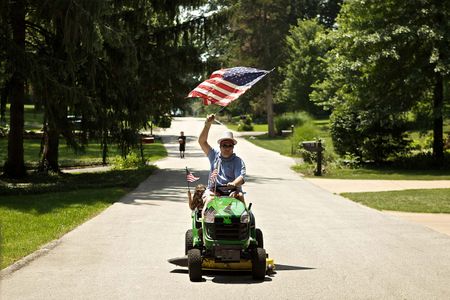 Patriotic man on John Deere tractor lawn mower driving down the street waving the American flag on July 4th, 4th of July. Man with American Flag on a Tractor