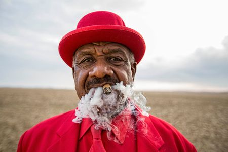 Portrait of William Seaberry, AKA Po Monkey staring into the camera smoking a cigar as cigar smoke billows up around his face. He is wearing a red suit standing in a field on a planation next to his juke joint. This photo reminds me of Sant Claus, as the smoke looks like a scant style beard:) Shot for the Mississippi Tourism campaign for advertising, branding, collateral and broadcast motion assets. Environmental portrait, and we are a industrial photographer, on location photographer & location commercial photographer. Shallow depth of field and selective focus. VisitMS William Seaberry, AKA Po Monkey in Red Suit