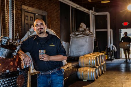 Environmental portrait of a man in his rum distillery holding a drink with a confident expression, in Vicksburg, MS. Shot for the Mississippi Tourism campaign for advertising, branding, collateral and broadcast motion assets. Environmental portrait, and we are a industrial photographer, on location photographer & location commercial photographer. VisitMS Man Sipping a Drink in a Distillery