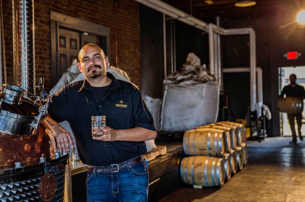 Environmental portrait of a man in his rum distillery holding a drink with a confident expression, in Vicksburg, MS. Shot for the Mississippi Tourism campaign for advertising, branding, collateral and broadcast motion assets. Environmental portrait, and we are a industrial photographer, on location photographer & location commercial photographer. VisitMS Man Sipping a Drink in a Distillery