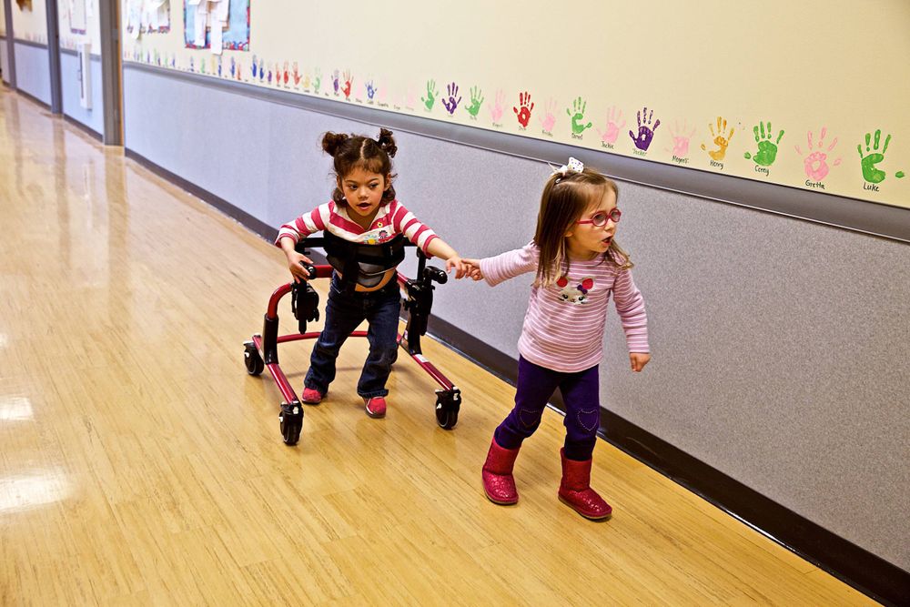 A photo of a two young girls in a school hallway holding hands, one of them is in a walker style wheelchair to help her walk, very affectionate, best friends, BFF. Photographed for a United Way advertising campaign. I am a advertising photographer, on location photographer & location commercial photographer. Photo of one of the many people that the United Way is assisting. Girls Walking in School Hallway