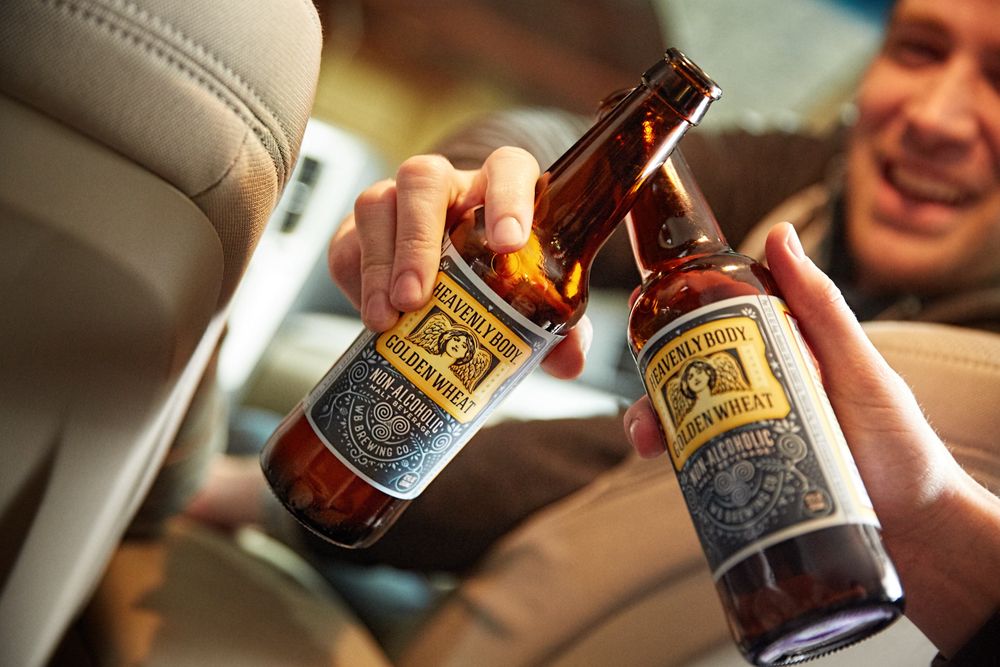 A couple of guys in a car going to lunch toasting & clinging a couple go bottles of WellBeing na non-alcoholic beers. Taosting in a Car