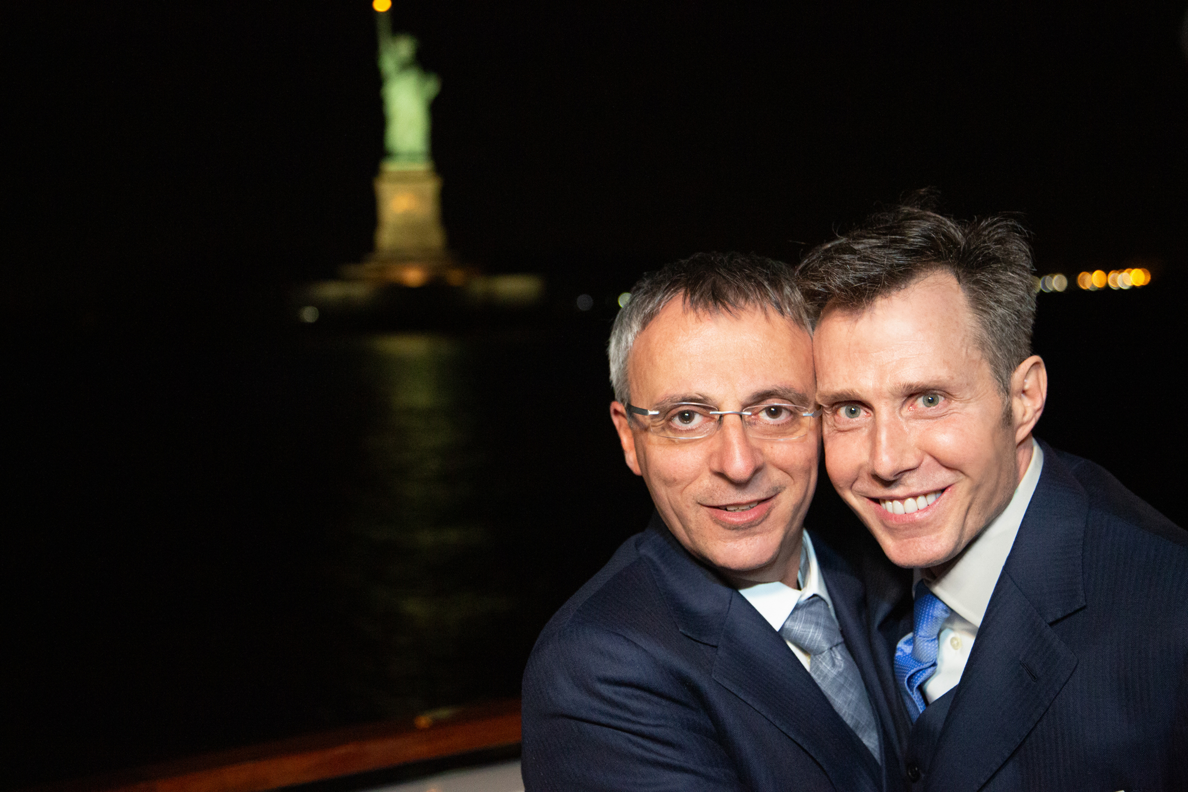 Paulo and Gianni cheek‑to‑cheek with Statue of Liberty behind them