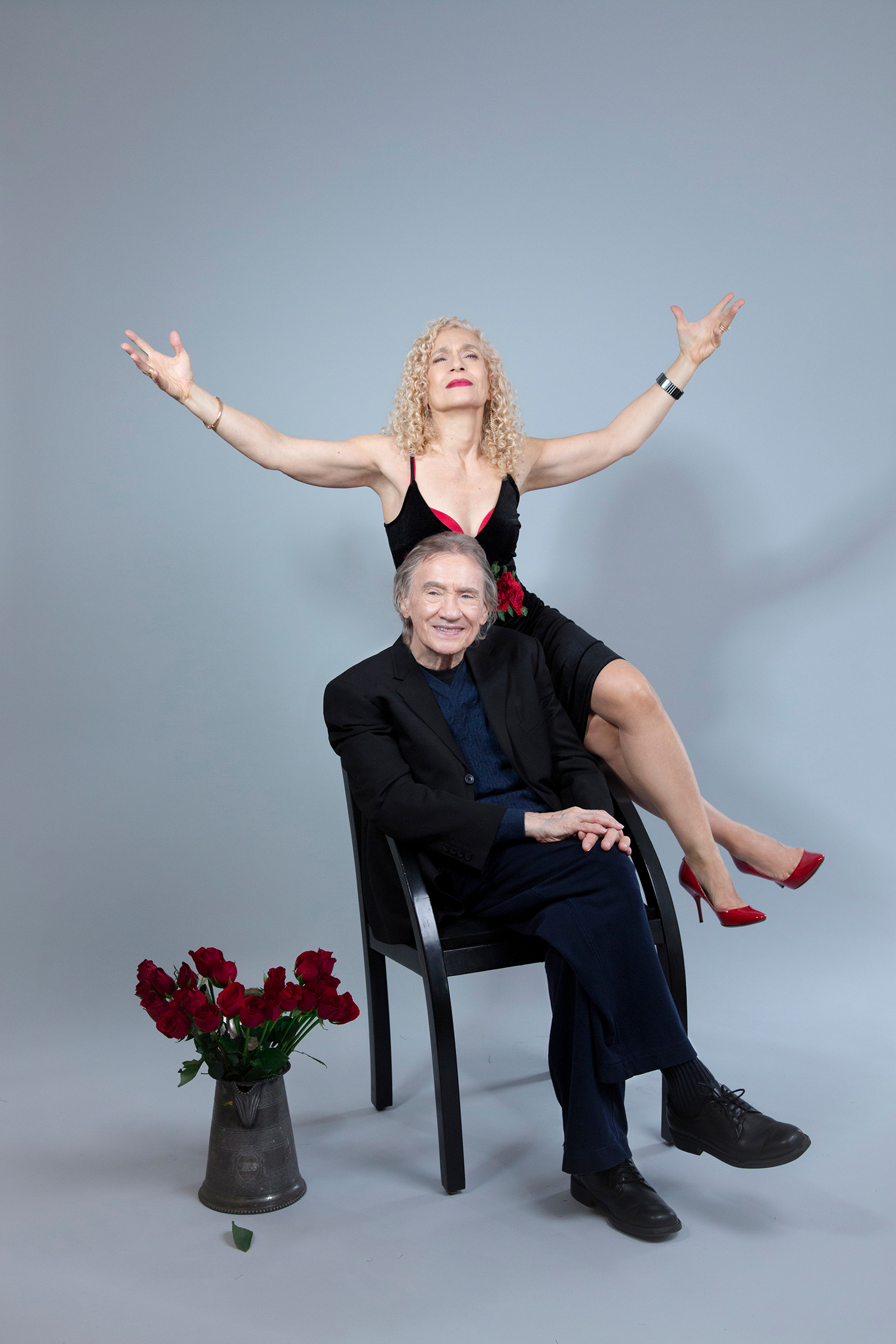 Irini singing with crossed legs behind Chuck in stylized long‑shot portrait