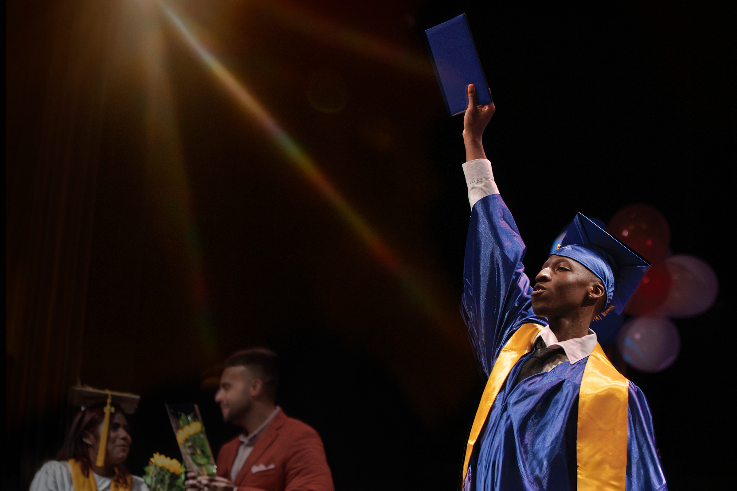 Black student raising diploma onstage with lens flare