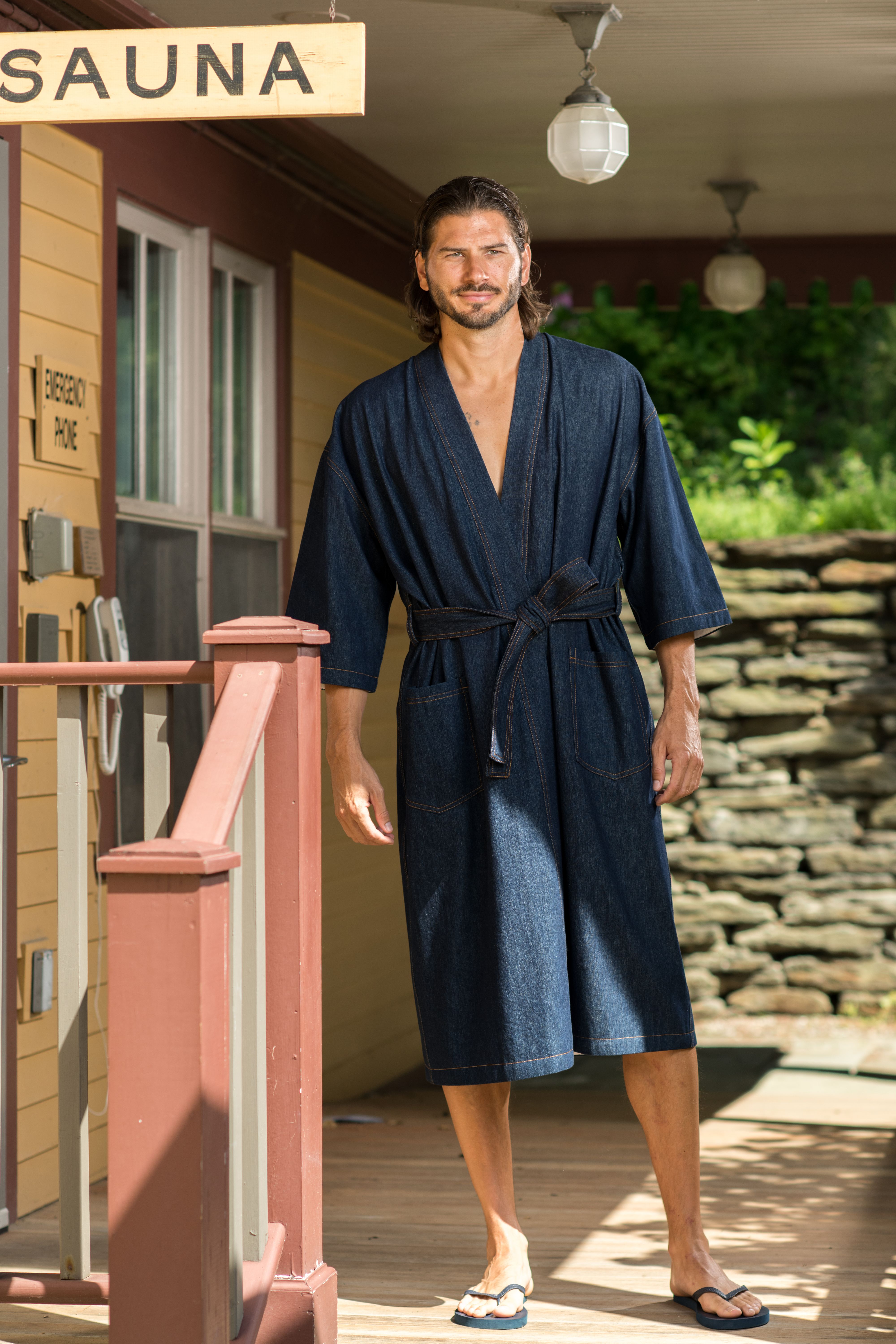 Male model in blue bathrobe stands on porch outside sauna