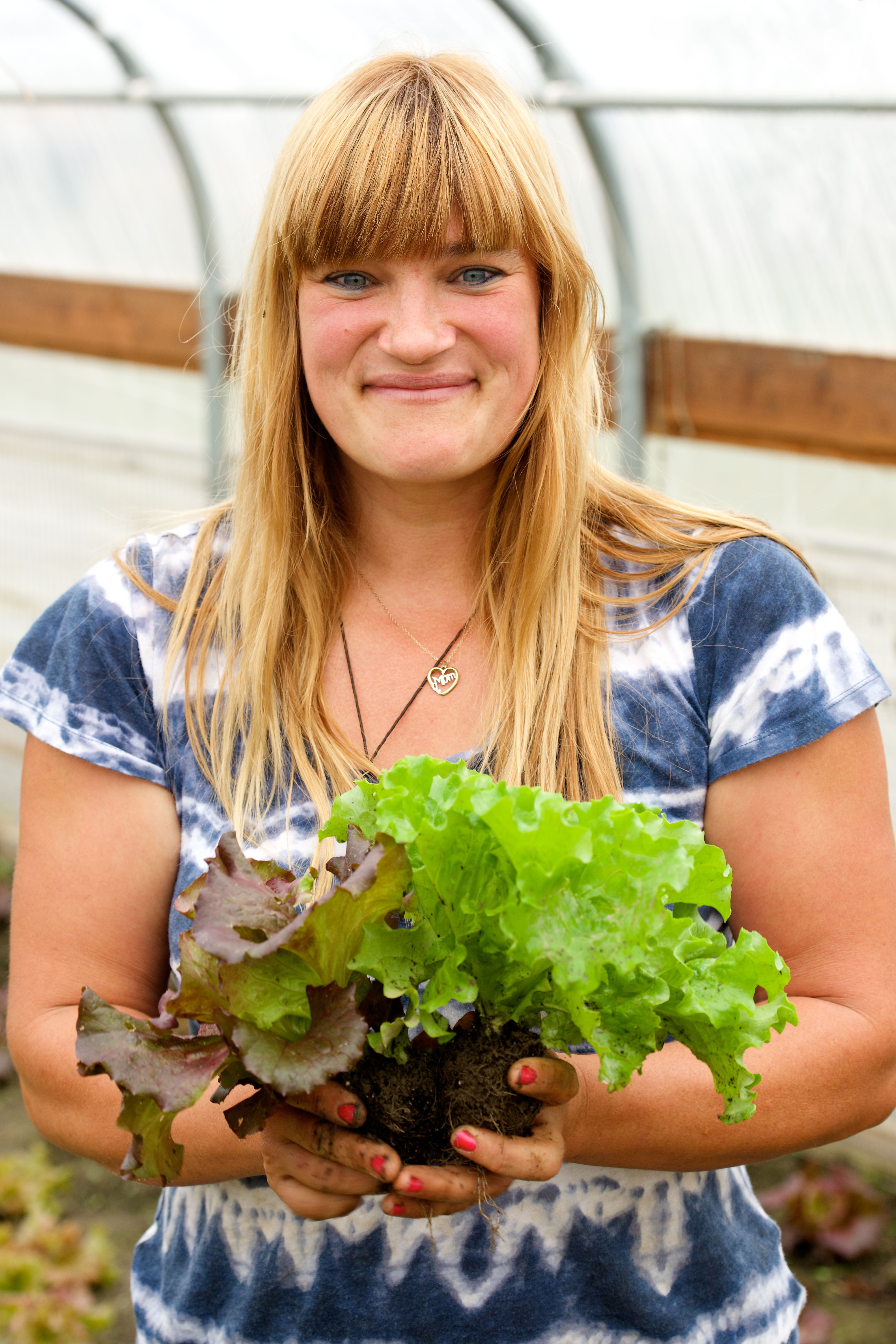 Blonde woman wearing blue tie die holds head of lettuce in greenhouse in lifestyle portrait
