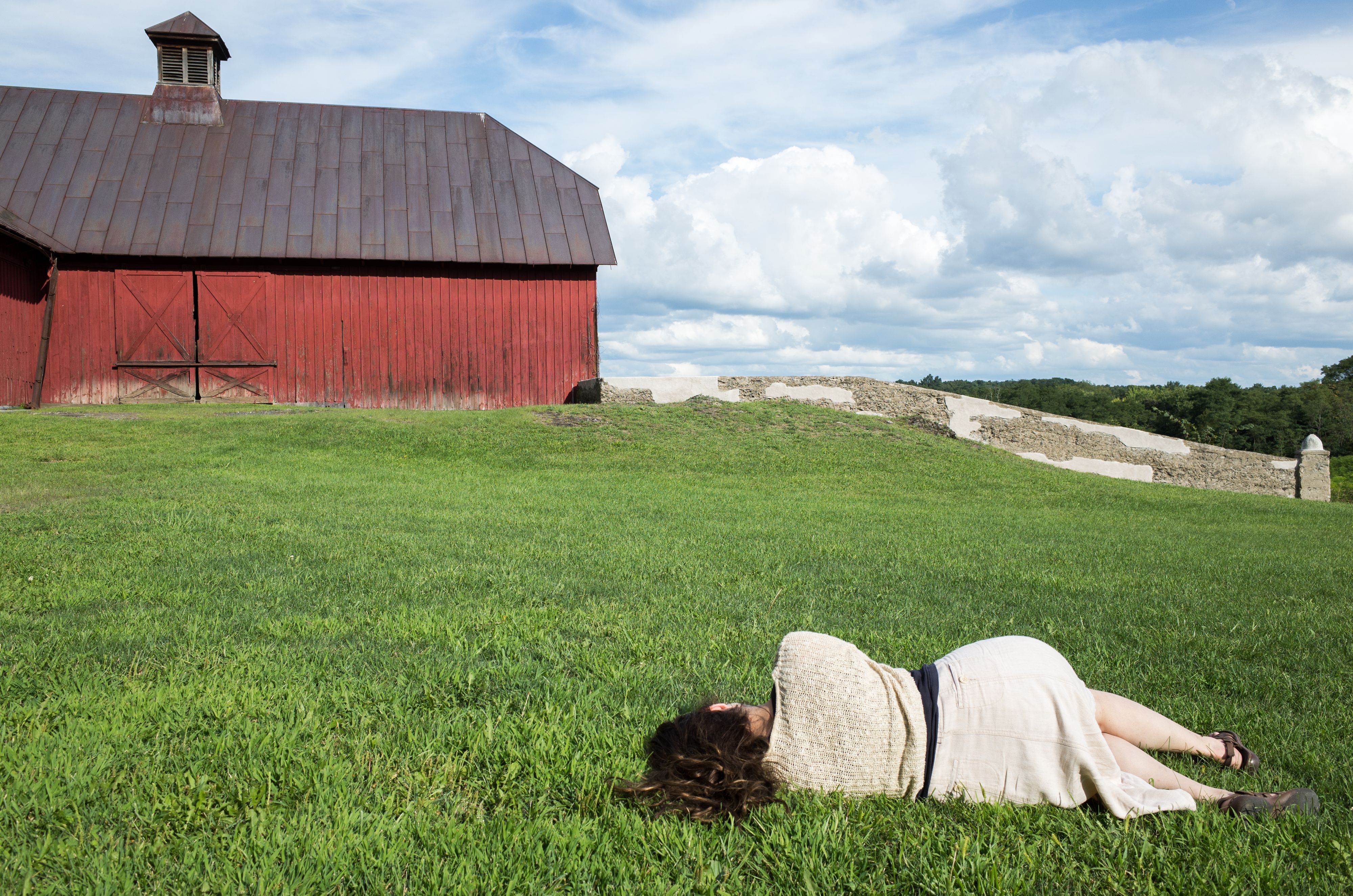 Woman in linen skirt lies in green field looking at red barn