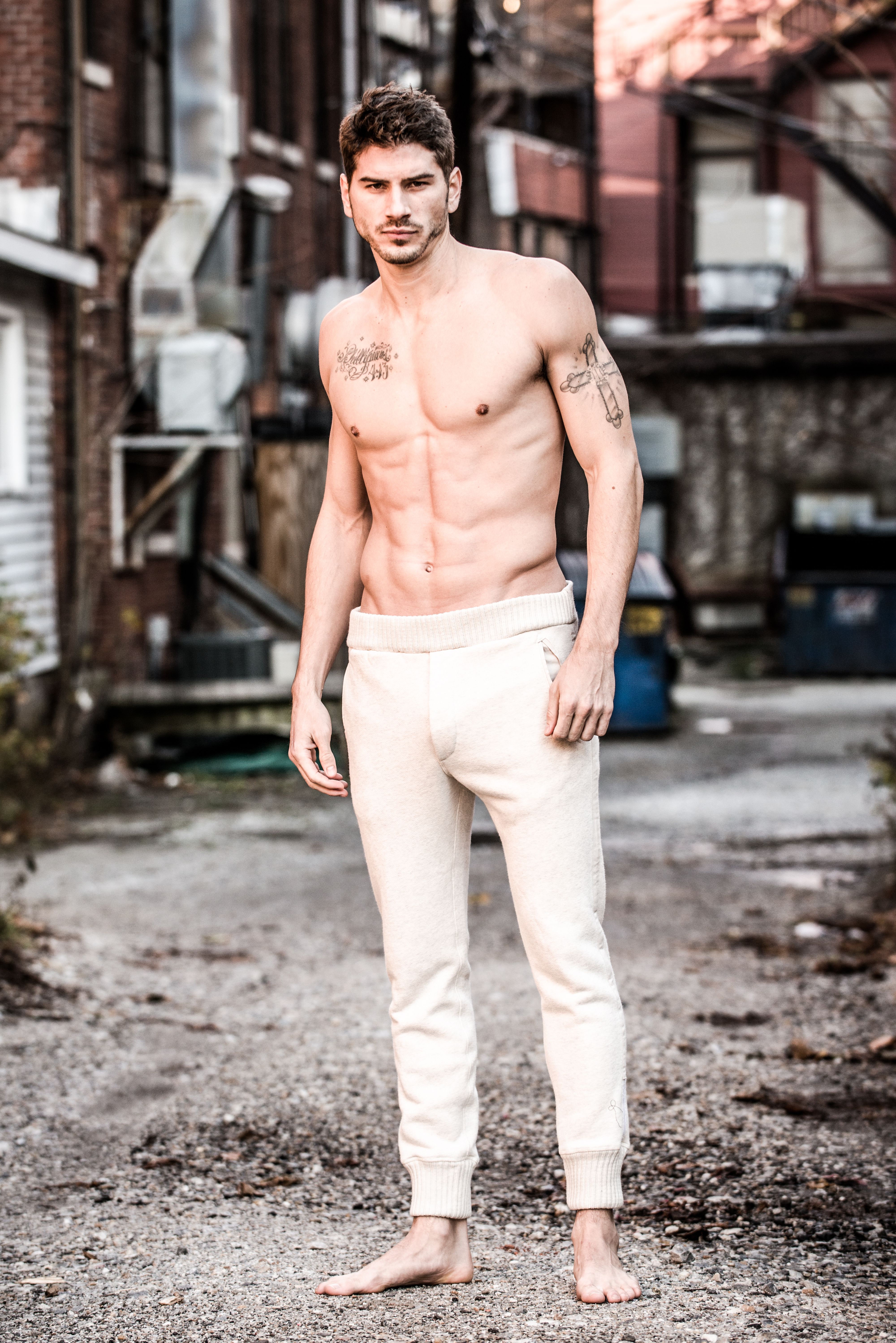 Shirtless and shoeless male model with tattoos and white pants standing in alleyway
