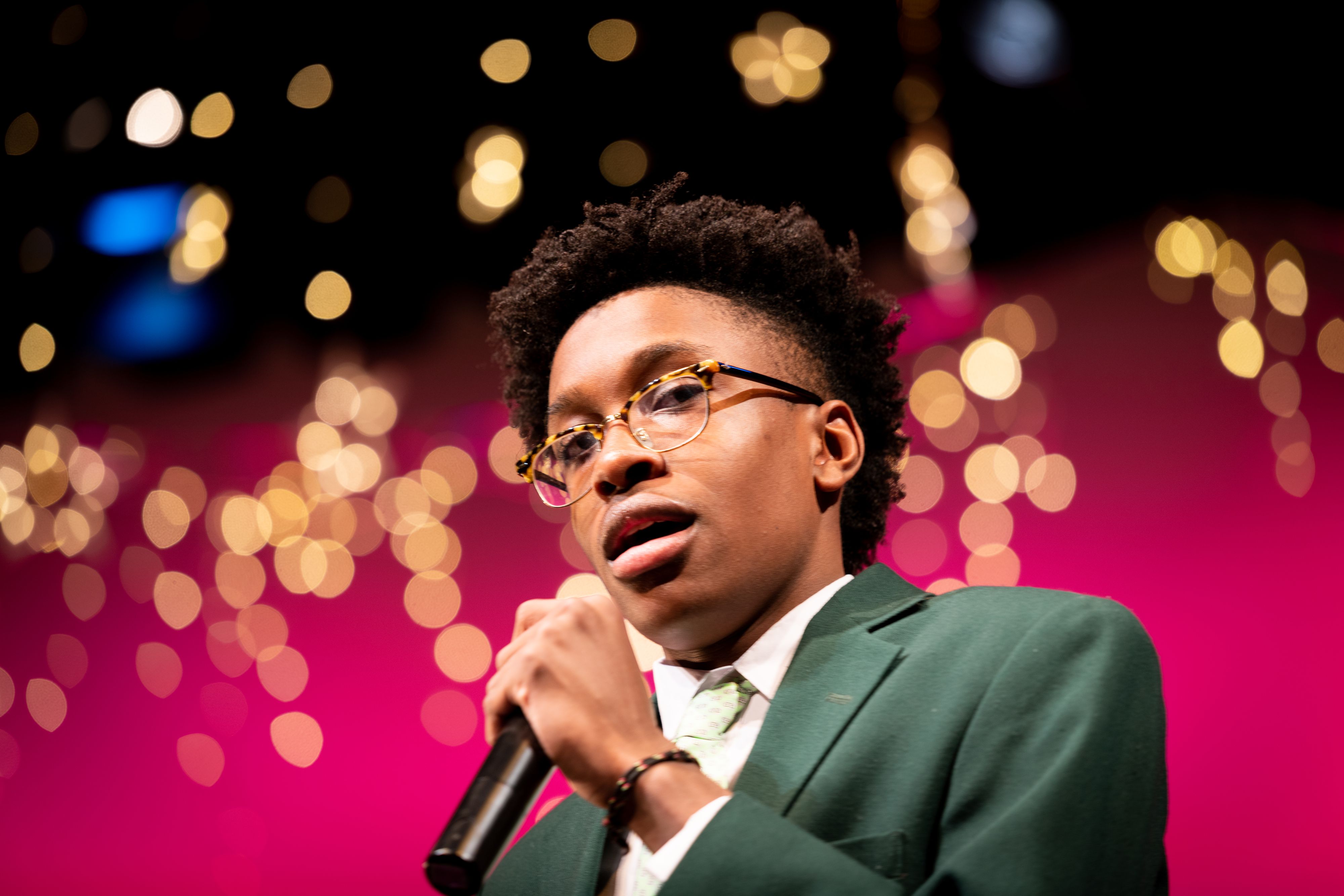 Young male African American in green jacket and tie singing with microphone looks at camera