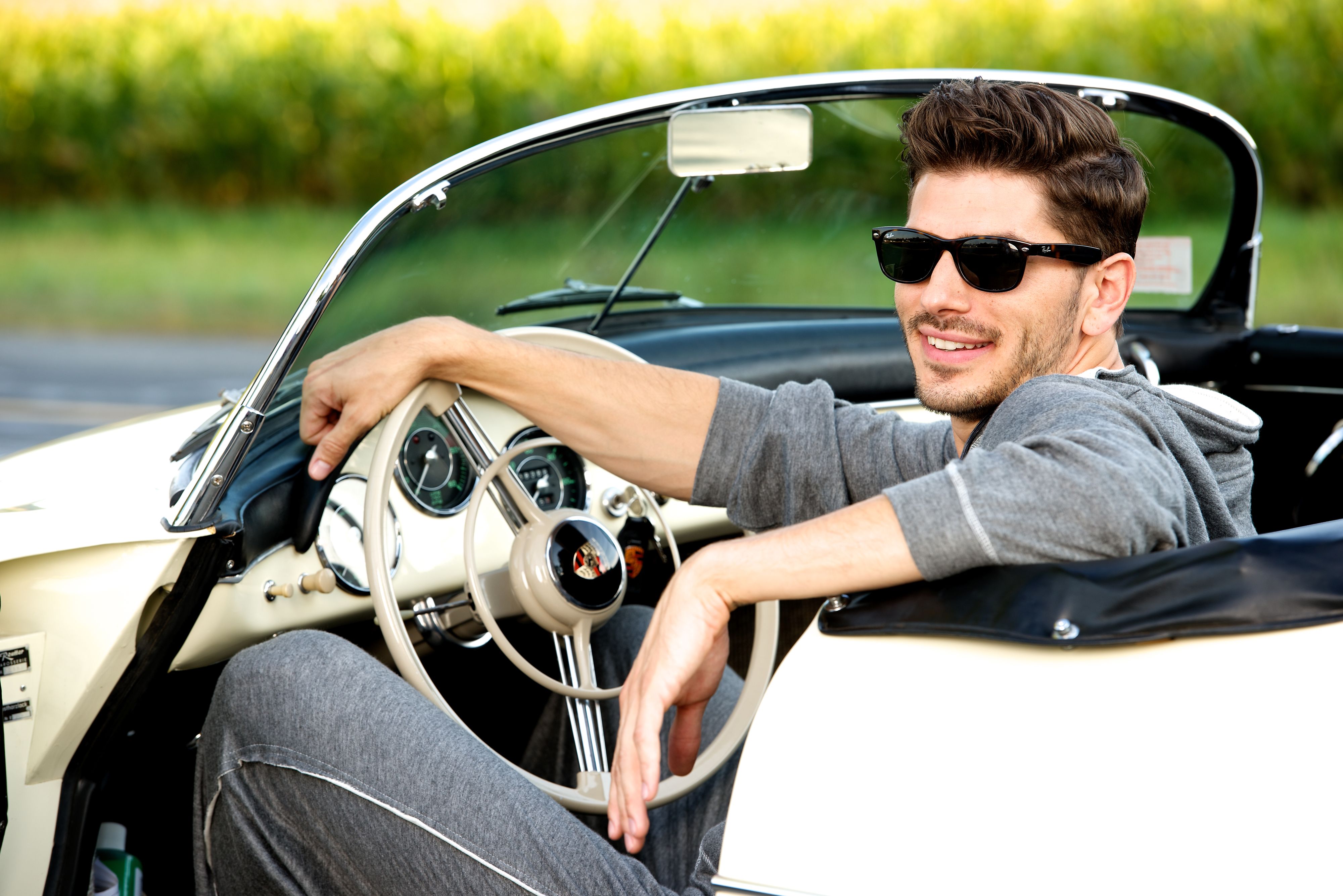 Male model wearing grey outfit and Wayfarer sunglasses sits in white Porsche 356 car
