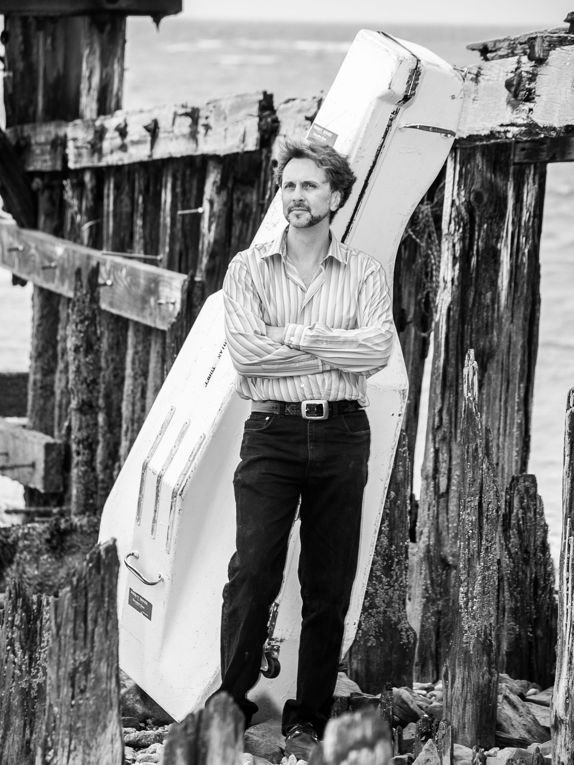 B&W portrait of cellist standing outside on pier with cello in shipping case
