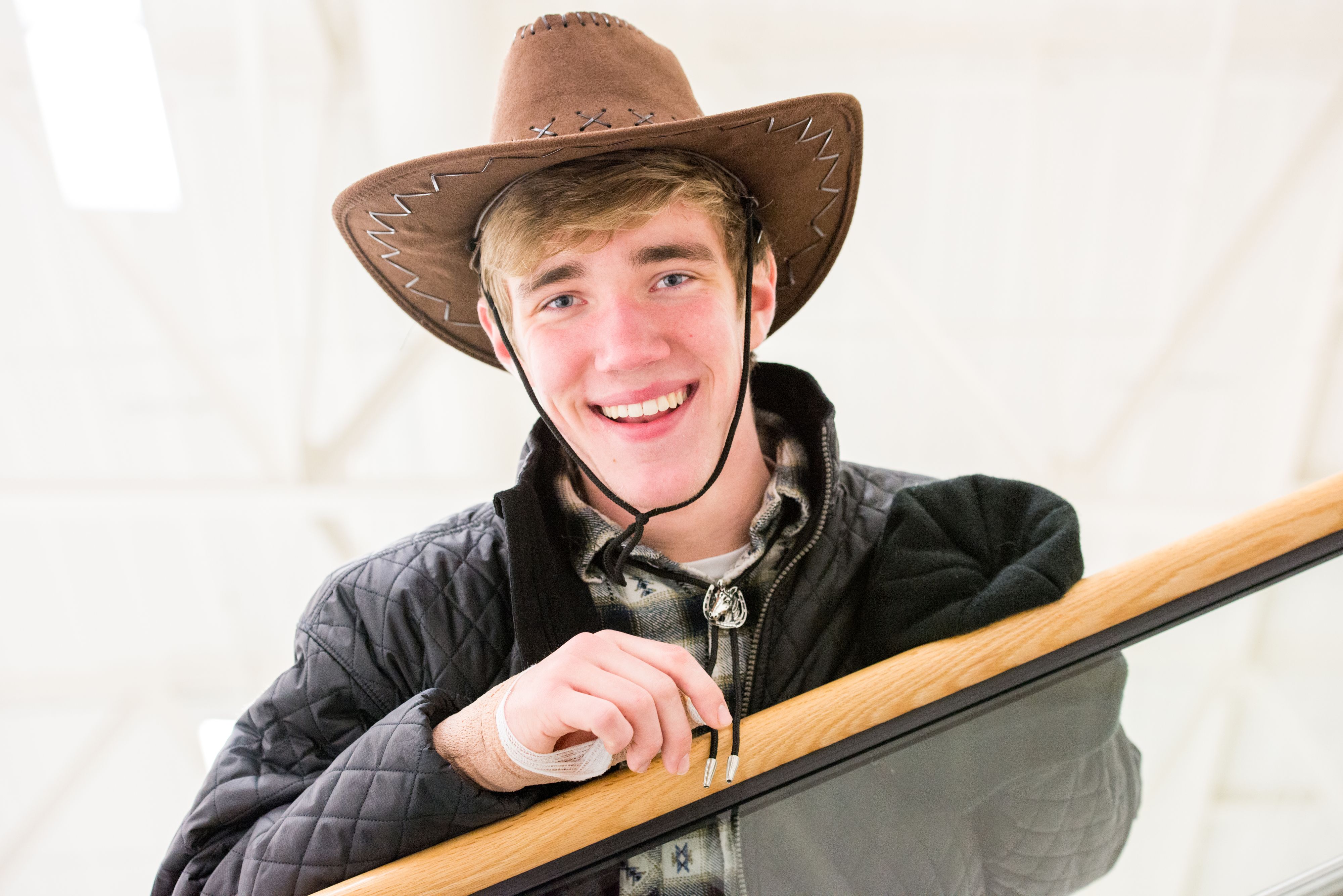 Male high school student wearing black quilted jacket and cowboy hat leaning on railing with smile
