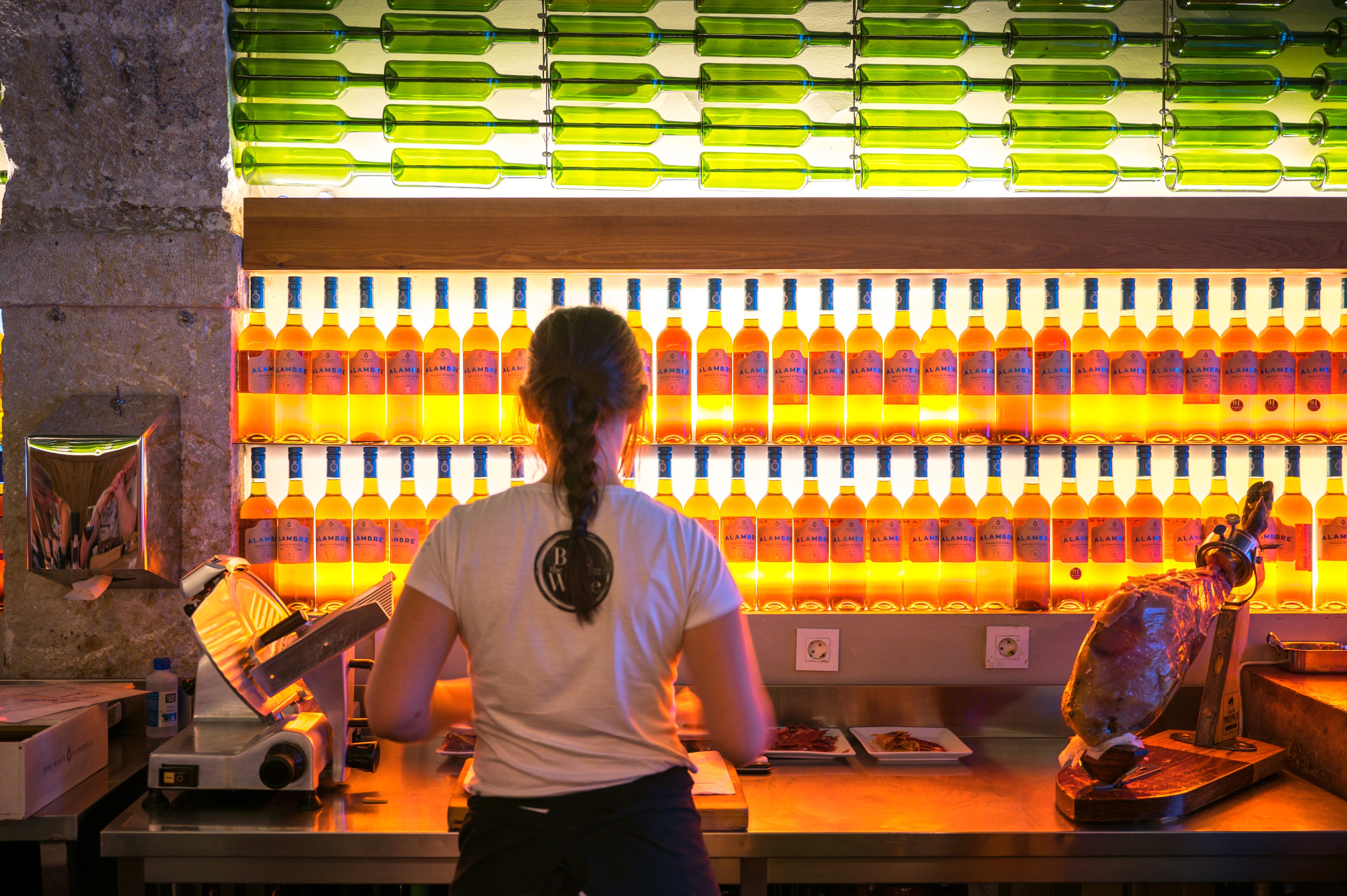 Server stands with back to camera and colorful backlit wine bottles covering wall