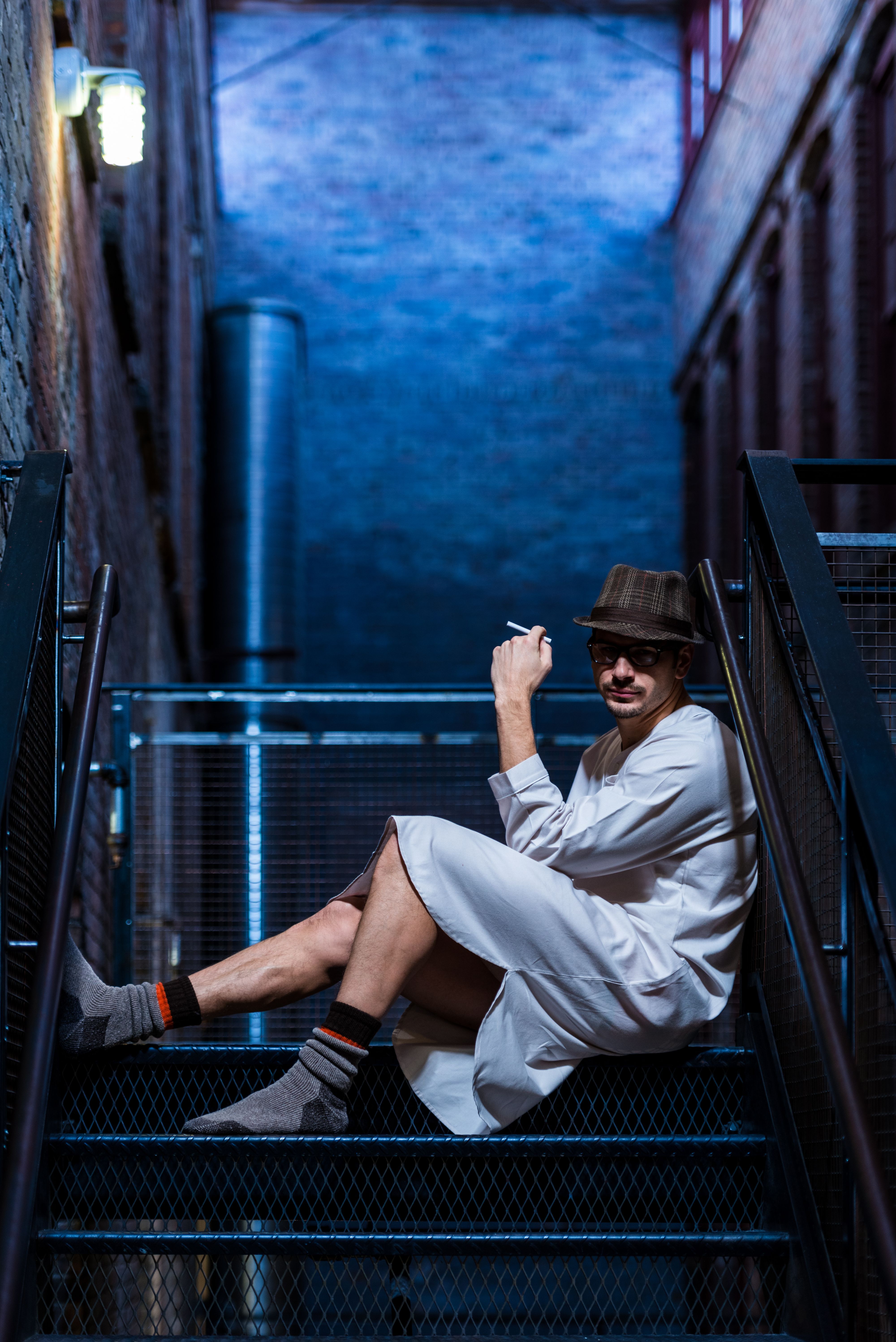 Male model in nightgown and hat sits on fire escape at night with cigarette