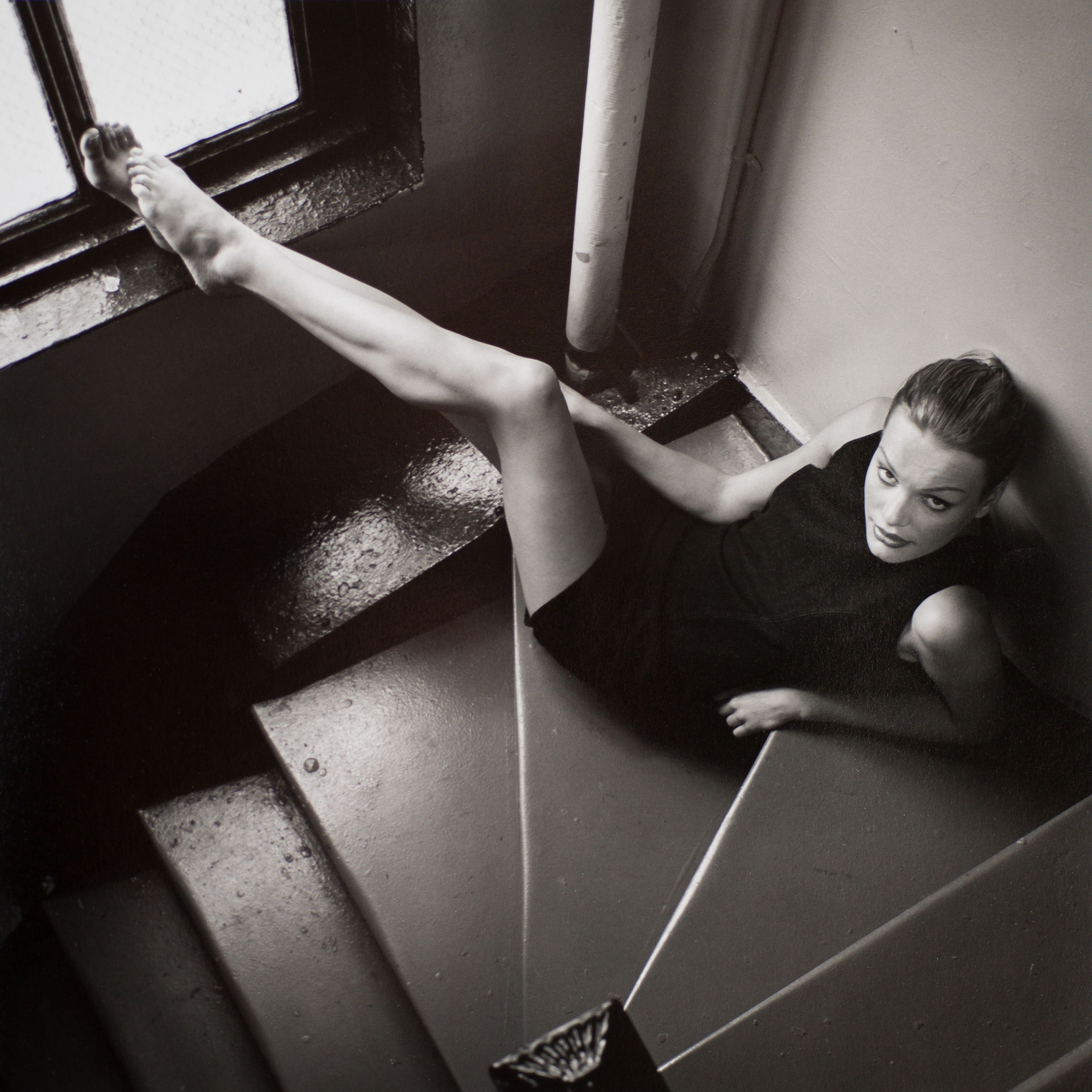 B&W photo of leggy model in black dress sitting in stairwell