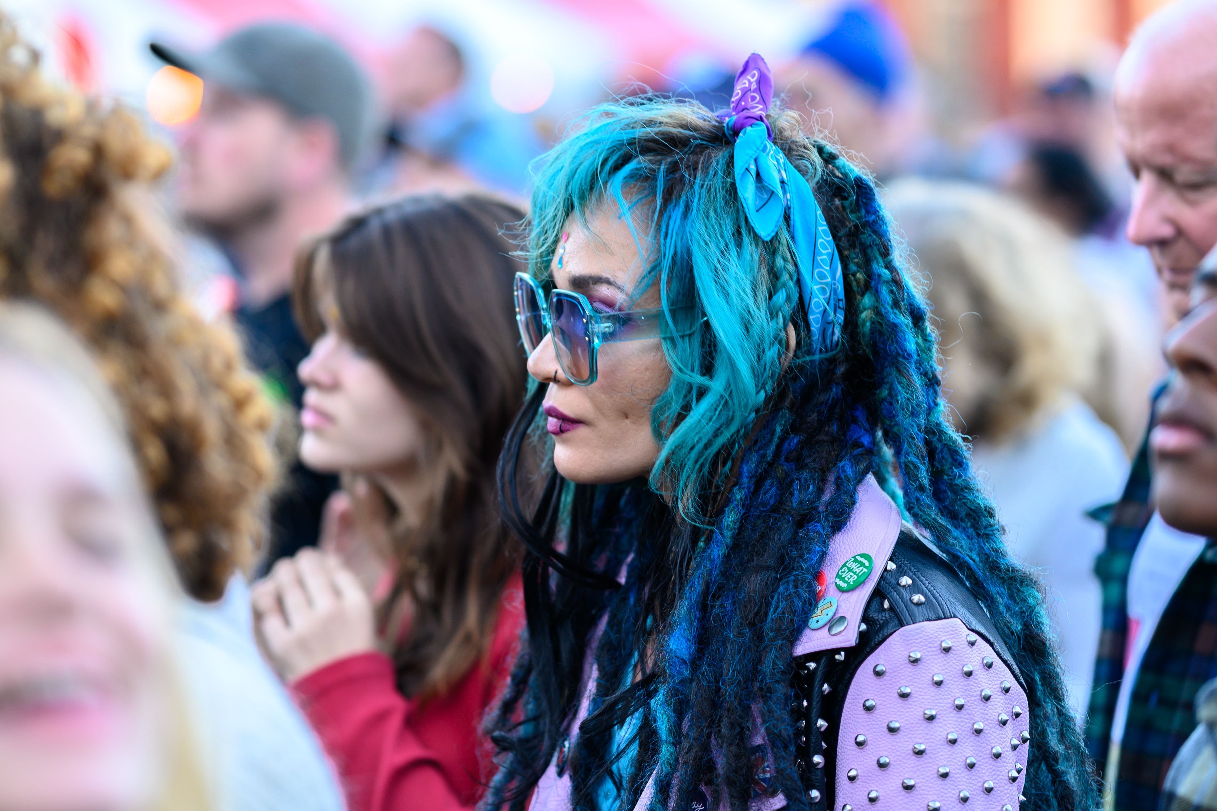 Woman with blue hair and glasses wearing pink leather jacket looks on at outdoor concert