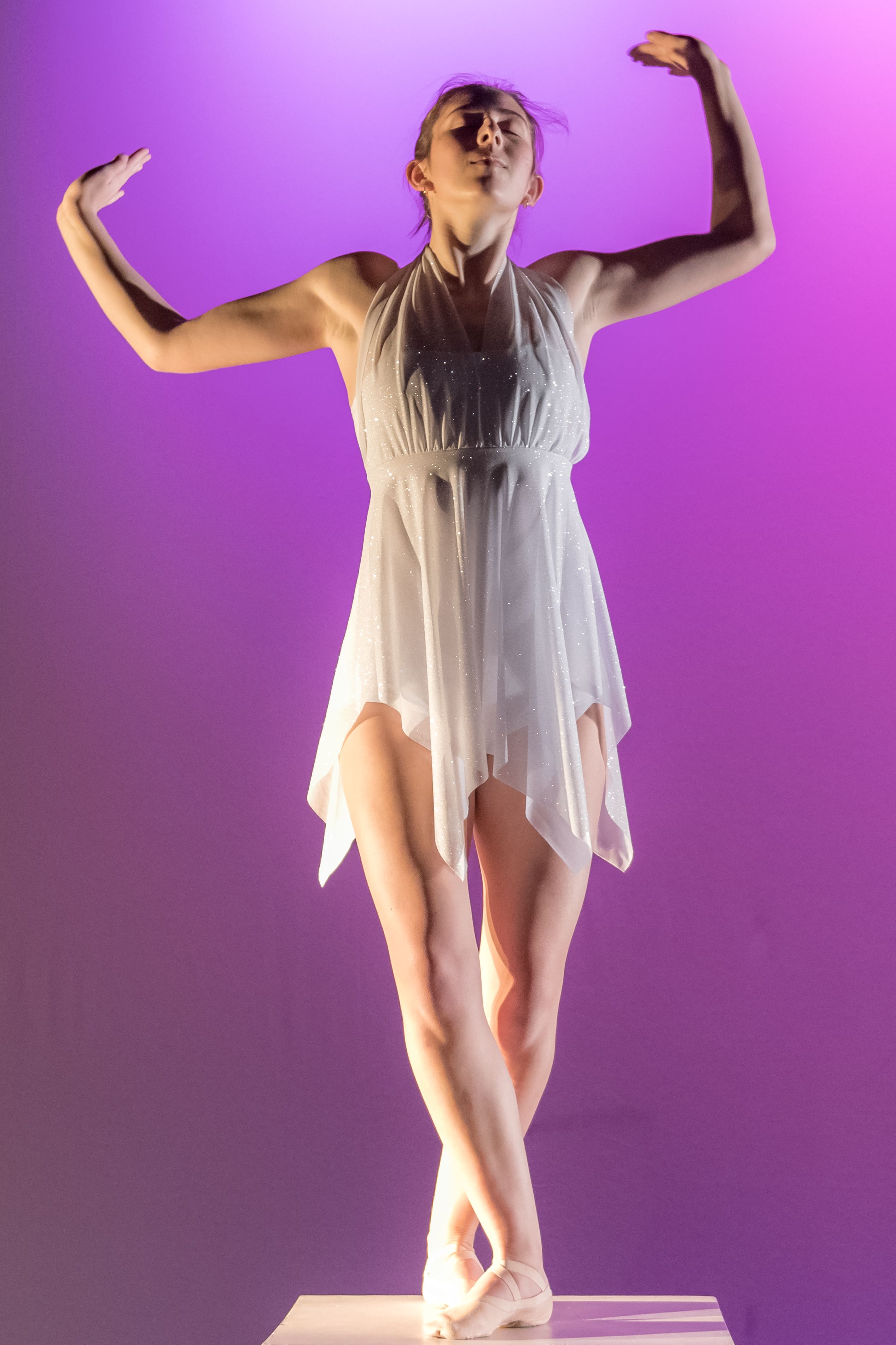 Female dancer with arms up and eyes closed on pedestal wearing white outfit on purple background