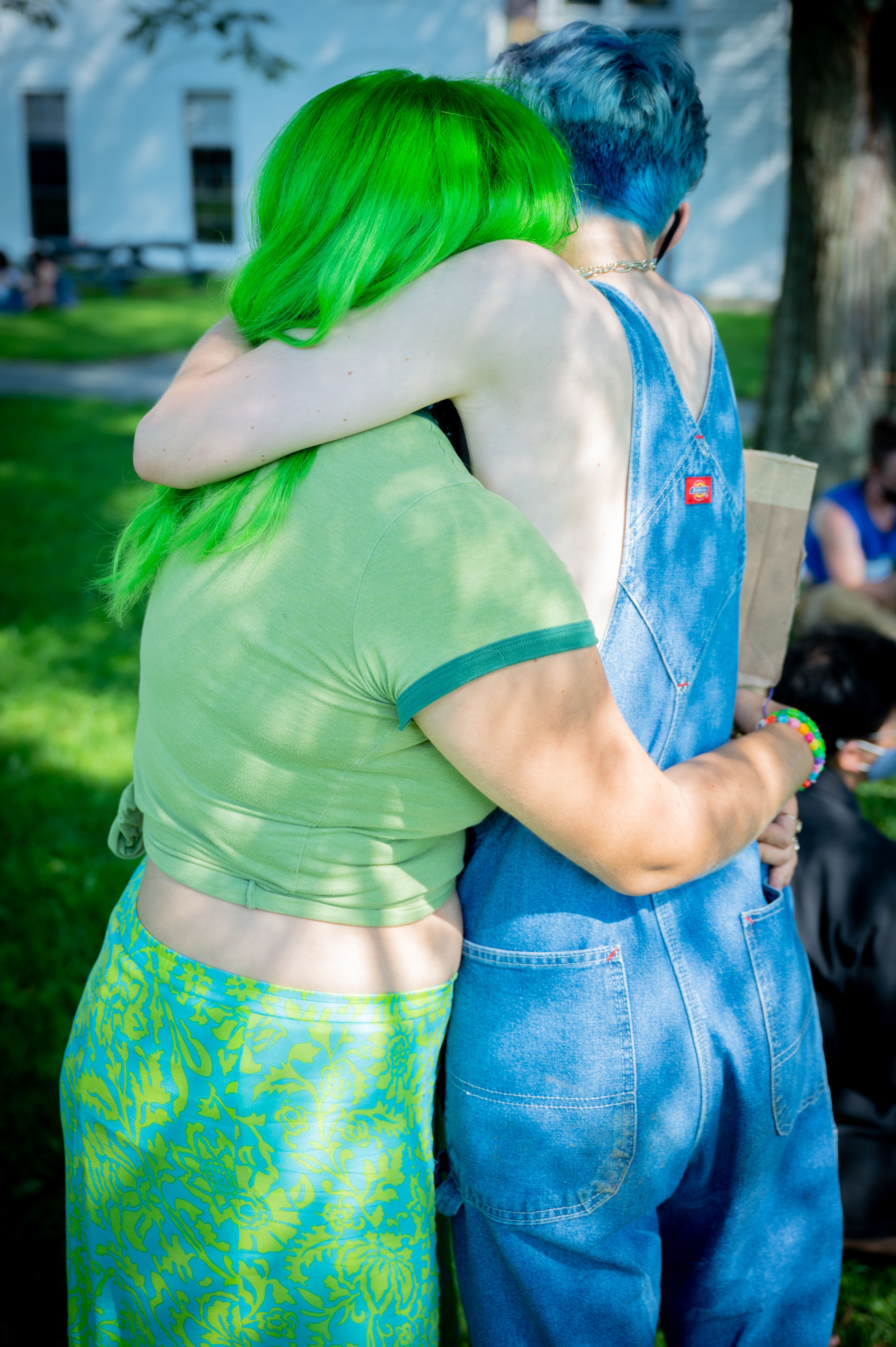 Two college student friends, one in green with green hair, the other in blue with blue hair hug with backs to camera
